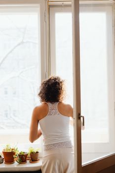 A woman with curly hair stands by an open window, surrounded by potted plants, enjoying a bright day.