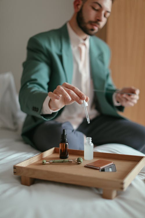 Elegantly Dressed Man Sitting on a Bed and Holding a Pipette Over a Bottle with a Cosmetic Product 