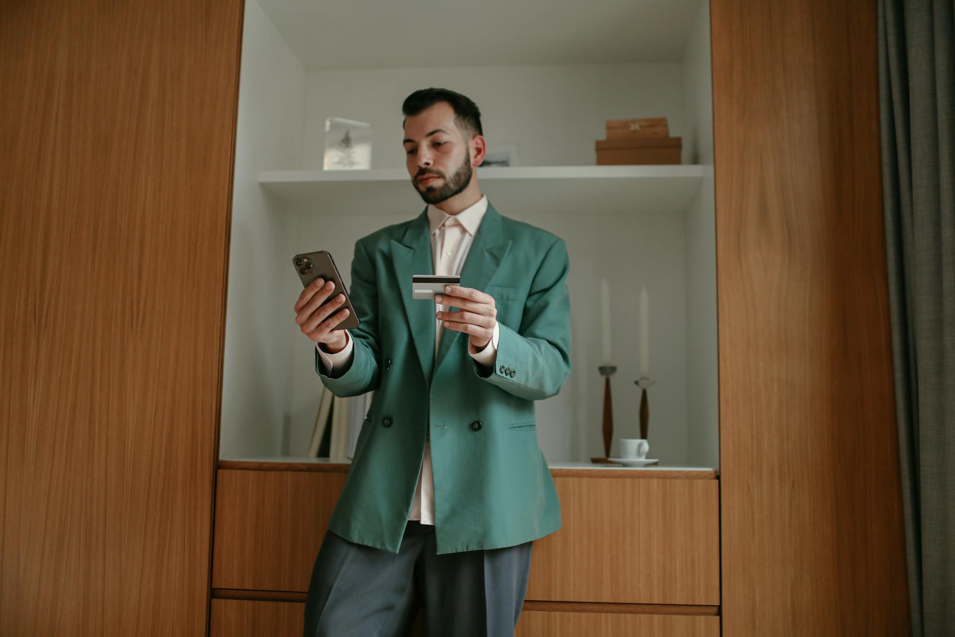 A bearded man in a green suit is using a smartphone and credit card indoors, showcasing modern technology and lifestyle.