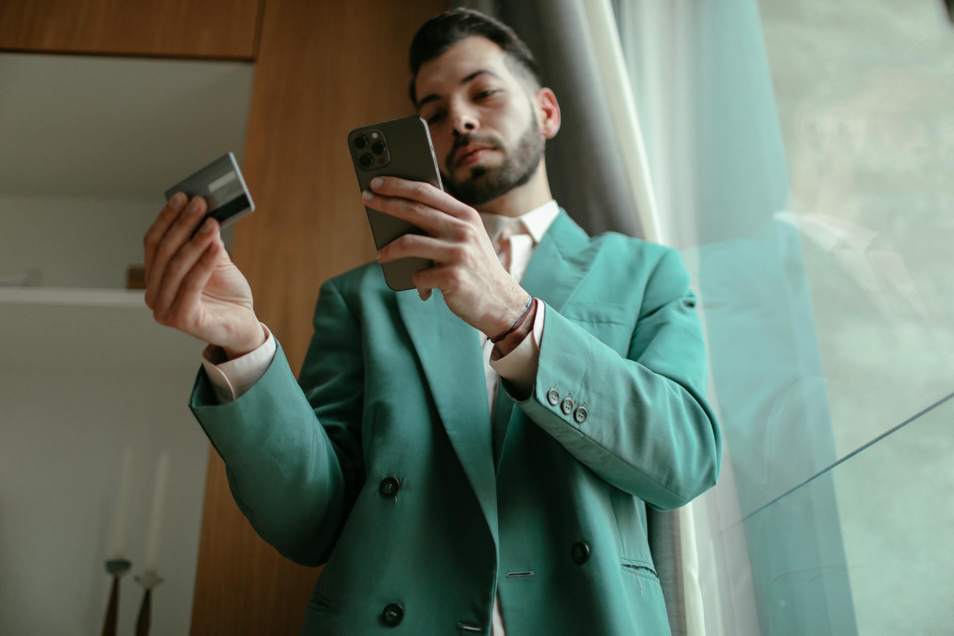 Man in teal blazer using smartphone and credit card for online shopping indoors.