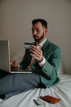 Adult man in teal blazer shopping online with credit card and laptop on bed.