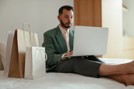 Adult man in teal blazer shopping online using laptop on bed with shopping bags.