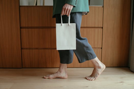 A person walks barefoot on wooden floor carrying a paper bag, dressed casually indoors.