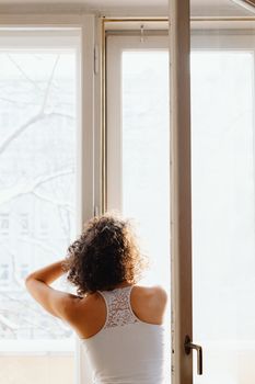 A woman stands in a sunlit room, gazing outside through an open window, enjoying the morning light.