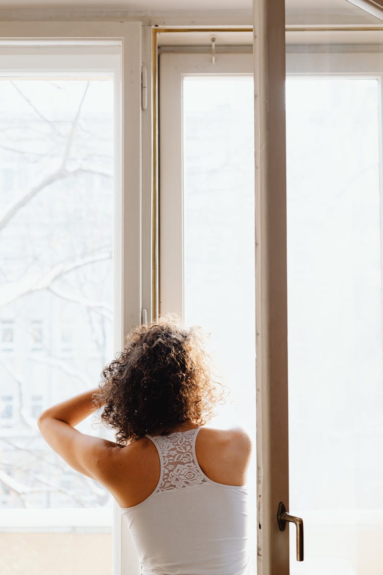 Woman Standing Next To An Open Window And Looking Outside