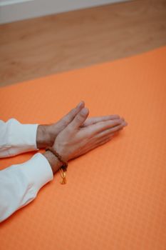 Close-up of hands with mala beads on an orange yoga mat indoors.