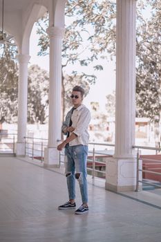 Young man in casual fashion wearing ripped jeans and sunglasses, standing stylishly outdoors.