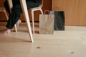 Side view of crop unrecognizable barefooted person resting on chair at table near paper bags placed on wooden floor at home