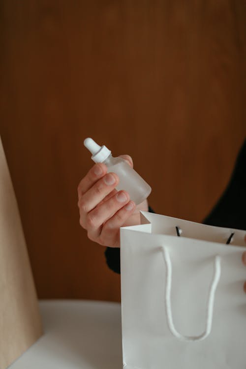 Crop faceless male buyer holding bottle of cosmetic serum sitting at table with shopping bags after receiving order