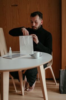 Full body of calm barefooted young guy sitting at table in modern apartment and checking purchases after successful shopping