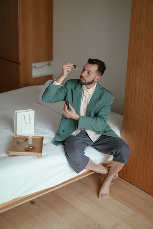 Trendy young guy applying cosmetic serum on face sitting on bed