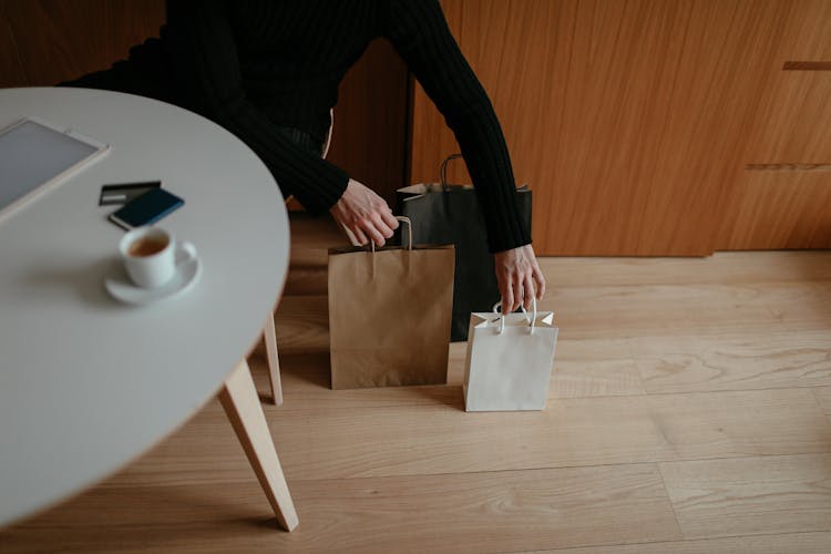 Anonymous Man Putting Shopping Bags On Floor Sitting At Table At Home