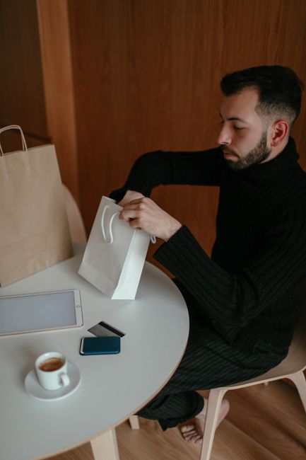 From above side view of serious barefooted young guy in casual clothes resting at table with cup of coffee and unpacking shopping bags From above side view of serious barefooted young guy in casual clothes resting at table with cup of coffee and unpacking shopping bags