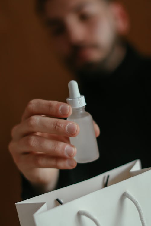 Crop unrecognizable young bearded male in black sweater demonstrating bottle of cosmetic facial moisturizing serum after shopping