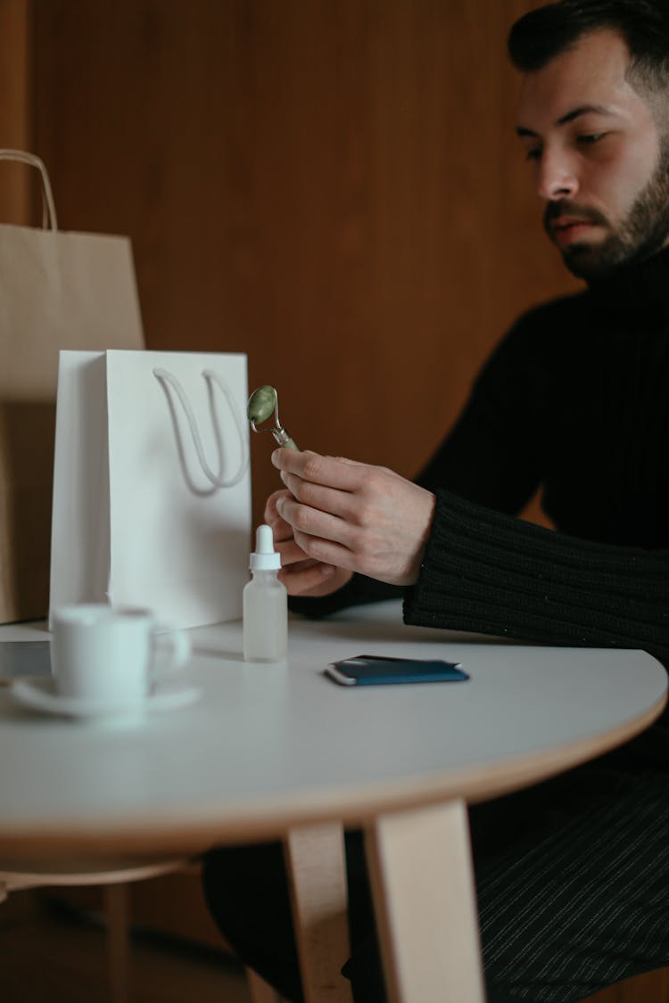 Metrosexual Young Guy Holding Jade Face Massager Sitting At Table At Home