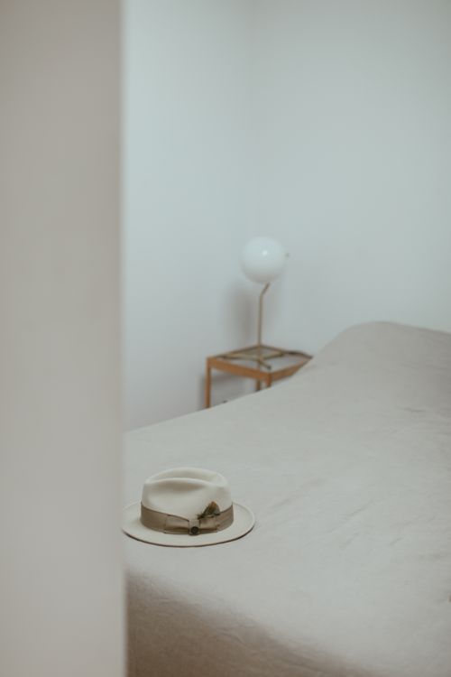 Free A serene minimalist bedroom with a fedora hat placed on a bed and a modern lamp on a side table. Stock Photo