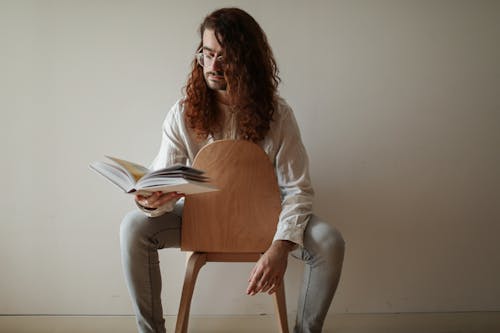 Free A Man Wearing White Long Sleeves Sitting on a Wooden Chair While Holding a Book Stock Photo