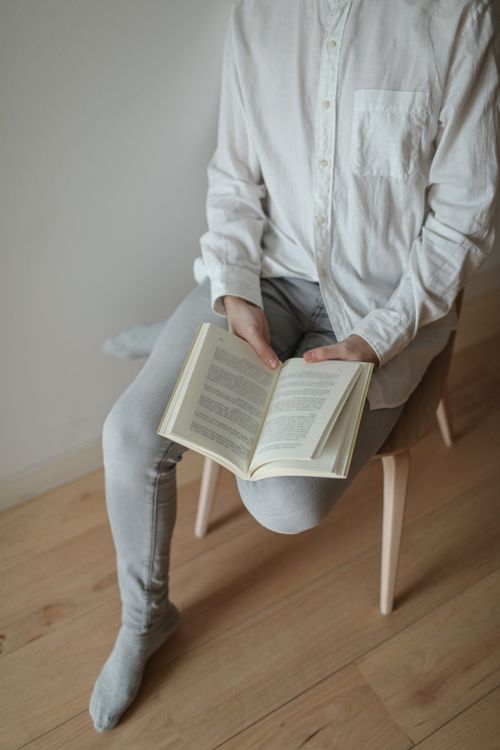 Free Adult reading a book while sitting on a wooden chair in a minimalist indoor setting. Stock Photo