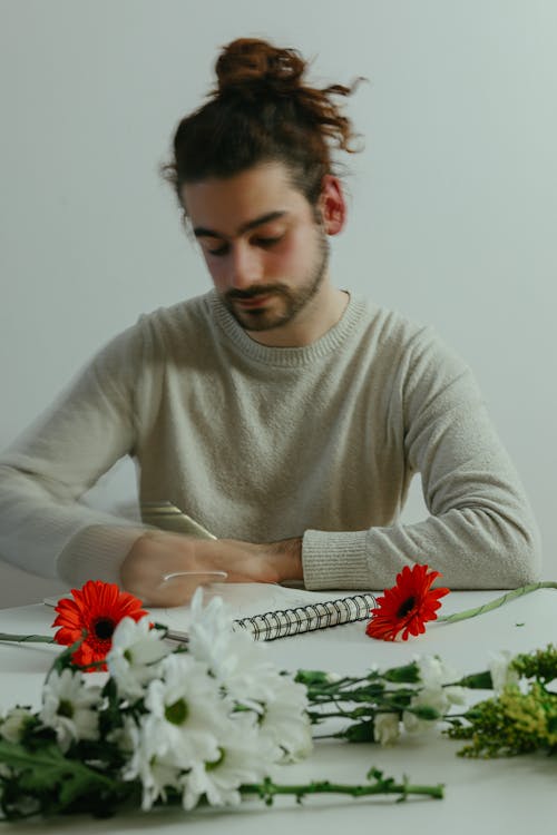 Free A Bearded Man in Beige Sweater Writing on Notebook on the Table with Flowers Stock Photo