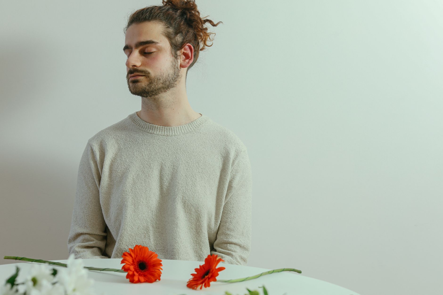 A man with facial hair and closed eyes sits at a white table with vibrant flowers, creating a serene mood.