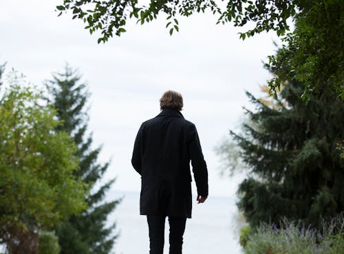 Fotografía De Enfoque Superficial Del Hombre Vestido Con Abrigo Negro Y Pantalones Negros De Pie Junto A árboles Verdes