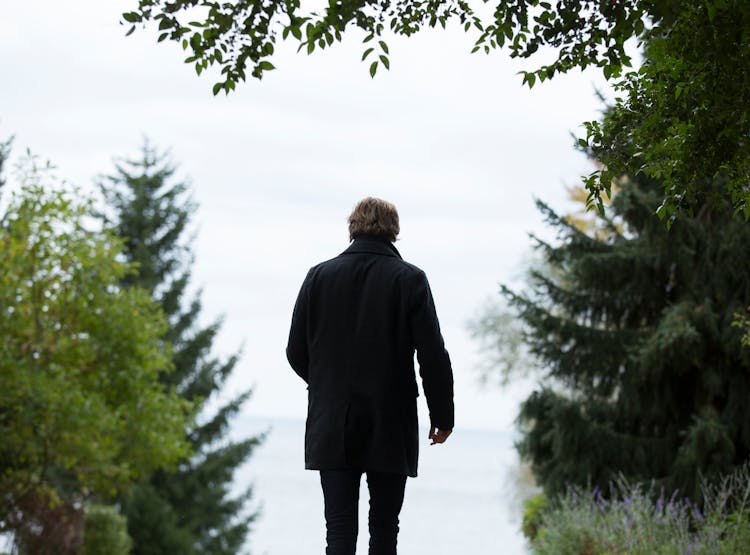 Shallow Focus Photography Of Man Wearing Black Coat And Black Pants Standing Beside Green Trees