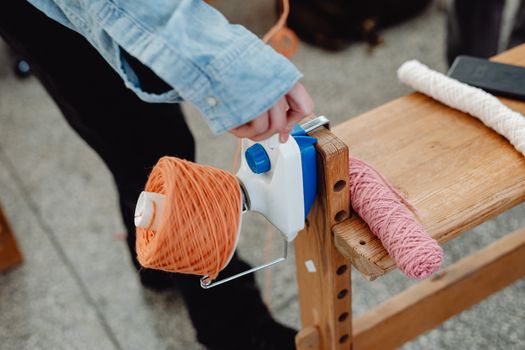 Close-up of a hand using a yarn winder with orange and pink thread skeins on a wooden table.