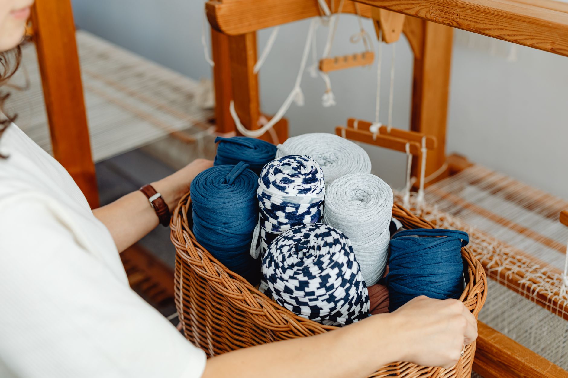 A woman's hands holding a basket of colorful fabric yarns near a wooden loom indoors.