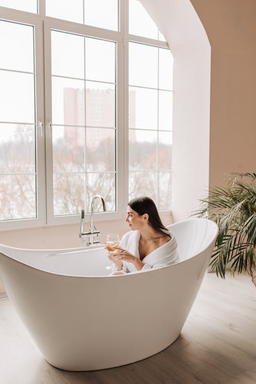 Woman in White Ceramic Bathtub Near Glass Window