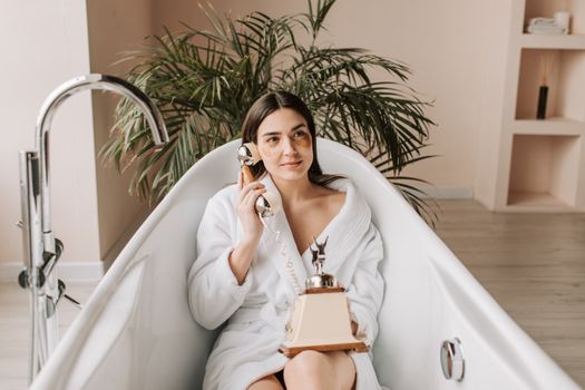 Serene woman in a bathrobe enjoying a call in a stylish bathtub indoors.