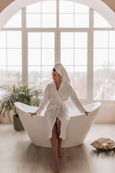 Woman in bathrobe and head towel sitting on bathtub edge, relaxing indoors.