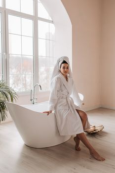 A sophisticated woman in a bathrobe sits gracefully by a modern bathtub in a bright, elegant bathroom.