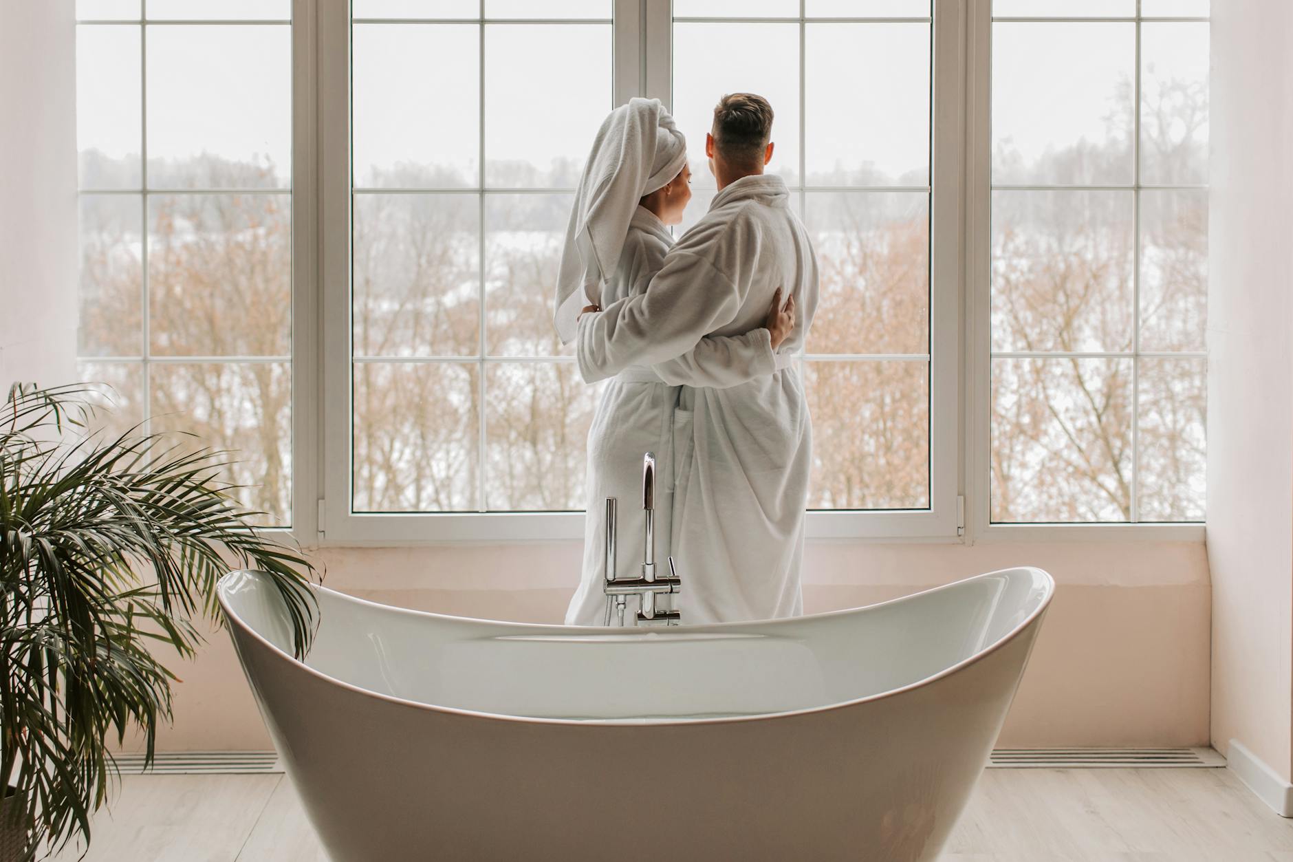 Couple in bathrobes standing by a window overlooking a snowy landscape.