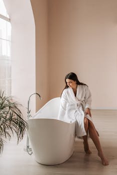 Serene woman in a white bathrobe sits by a modern bathtub in a minimalist bathroom.