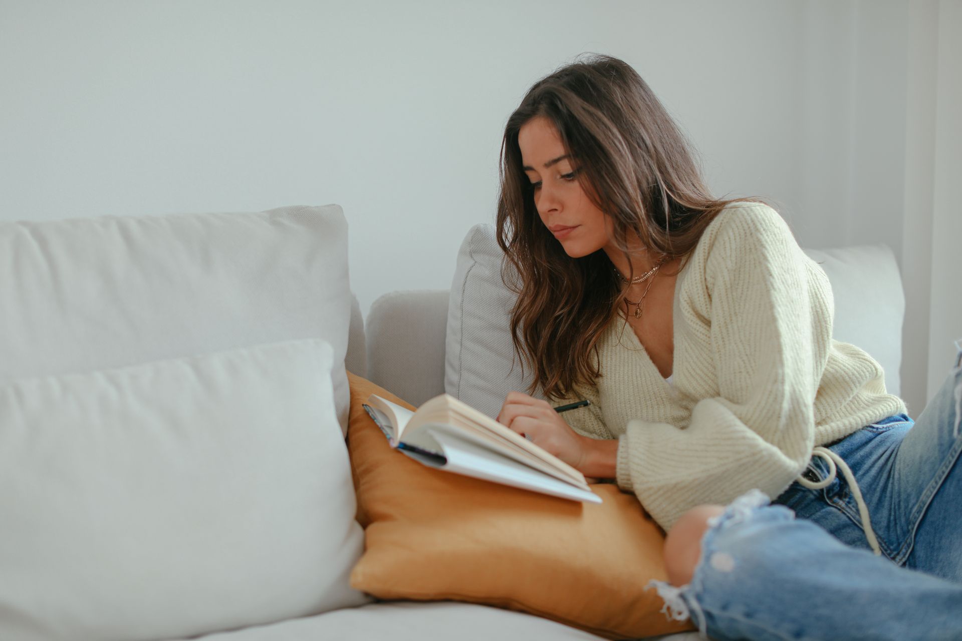 A young woman reclines on a couch reading a book in a cozy home setting.