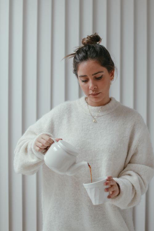 Free A woman in cozy attire pouring tea into a cup by a window, creating a warm and serene atmosphere. Stock Photo