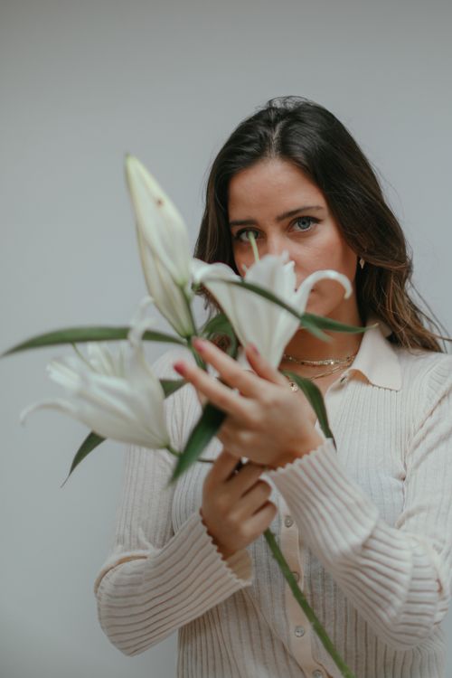 Free A serene woman in a sweater holding a white lily bouquet against a plain background. Stock Photo
