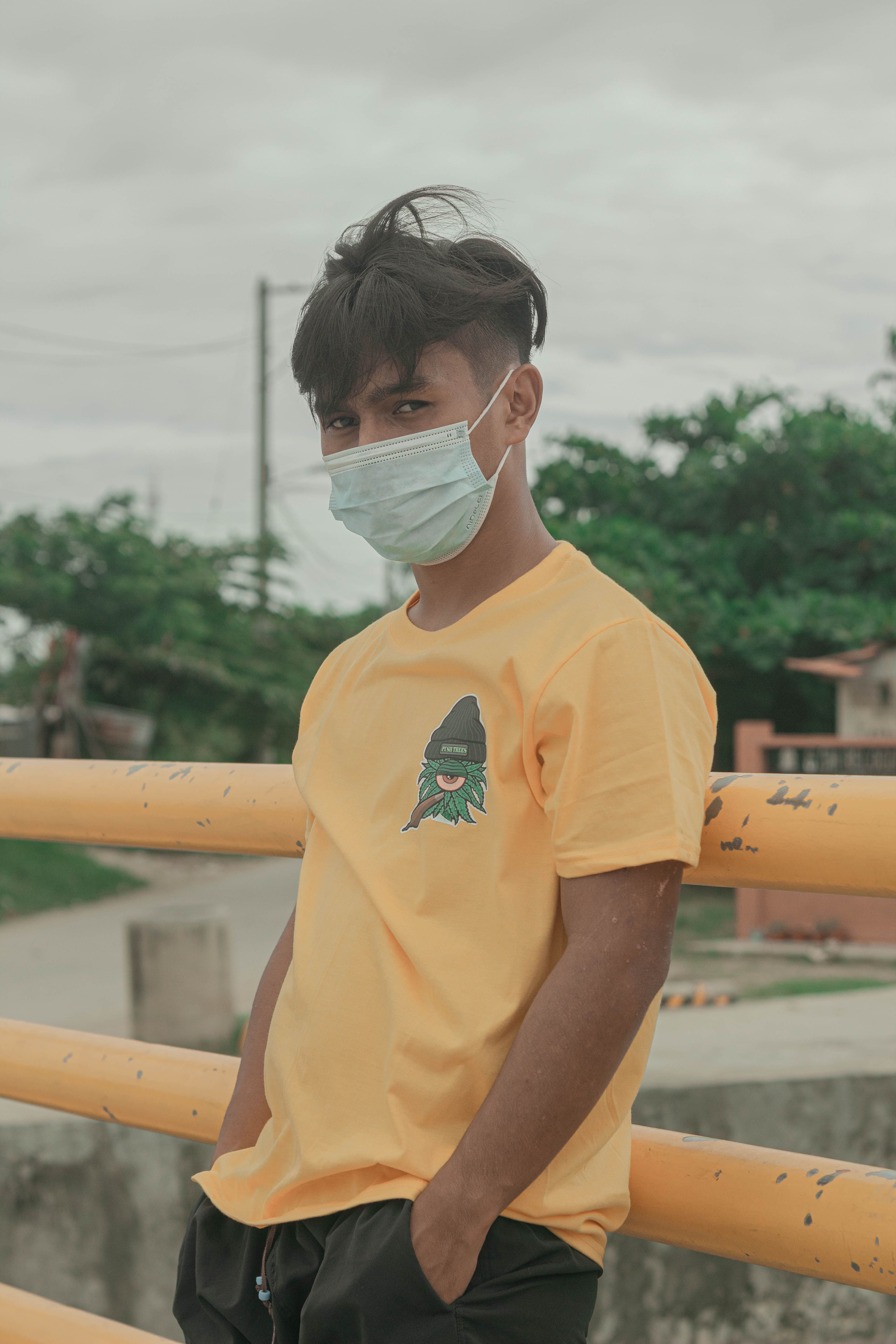 A young man wearing a mask and yellow shirt outdoors, adapting to new norms.
