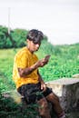 Man in Yellow Shirt Sitting on Concrete Bench while using Mobile Phone