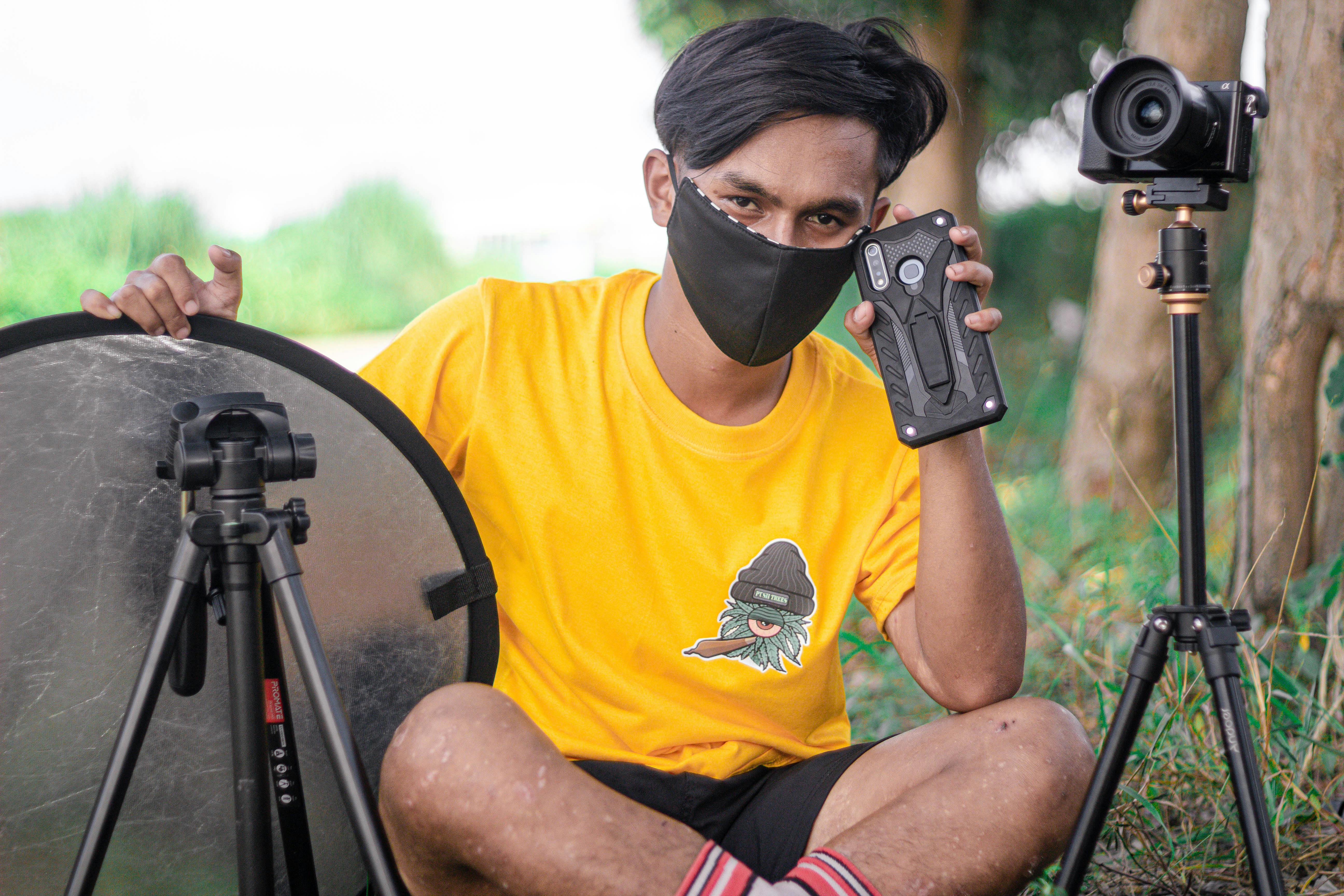 Photographer outdoors with DSLR camera, reflector, and smartphone. Wearing a yellow t-shirt and face mask.