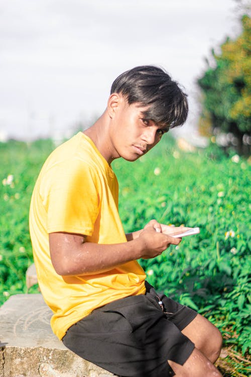 Portrait of a Man in Yellow T-Shirt Sitting a Green Field and Holding a Mobile Phone 