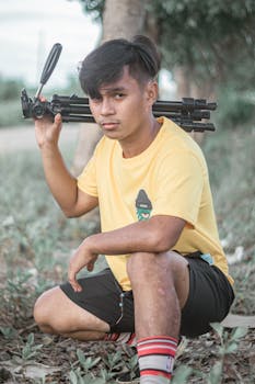 Casual portrait of a young man kneeling outdoors with a camera tripod on his shoulder.