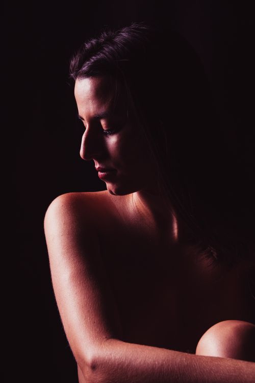 Free Graceful young naked ethnic female model embracing knees and looking down whil sitting in dark studio Stock Photo