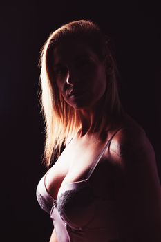 Alluring young female with blond hair in sexy lingerie looking at camera while standing in dark studio