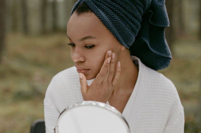 A Black woman with a navy towel wrapped on her hair and a white robe touches her cheek as she checks her skin in a small mirror.