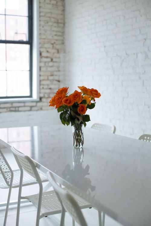 Free A bright, minimalist office space featuring a vase of vibrant orange roses on a sleek white table. Stock Photo