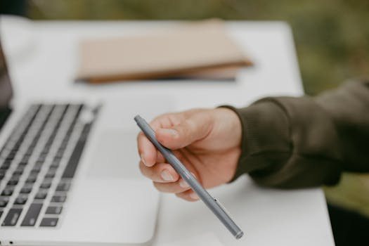 Close-up of a person holding a ballpoint pen near a laptop, symbolizing work or study.