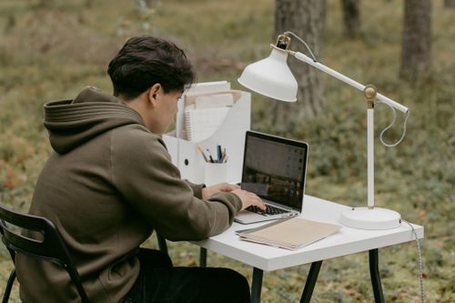 Free Man in hoodie using laptop at an outdoor workspace in a serene forest setting. Stock Photo