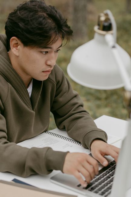 Young man in olive jacket working intently on a laptop while outdoors, with notebooks nearby. Young man in olive jacket working intently on a laptop while outdoors, with notebooks nearby.