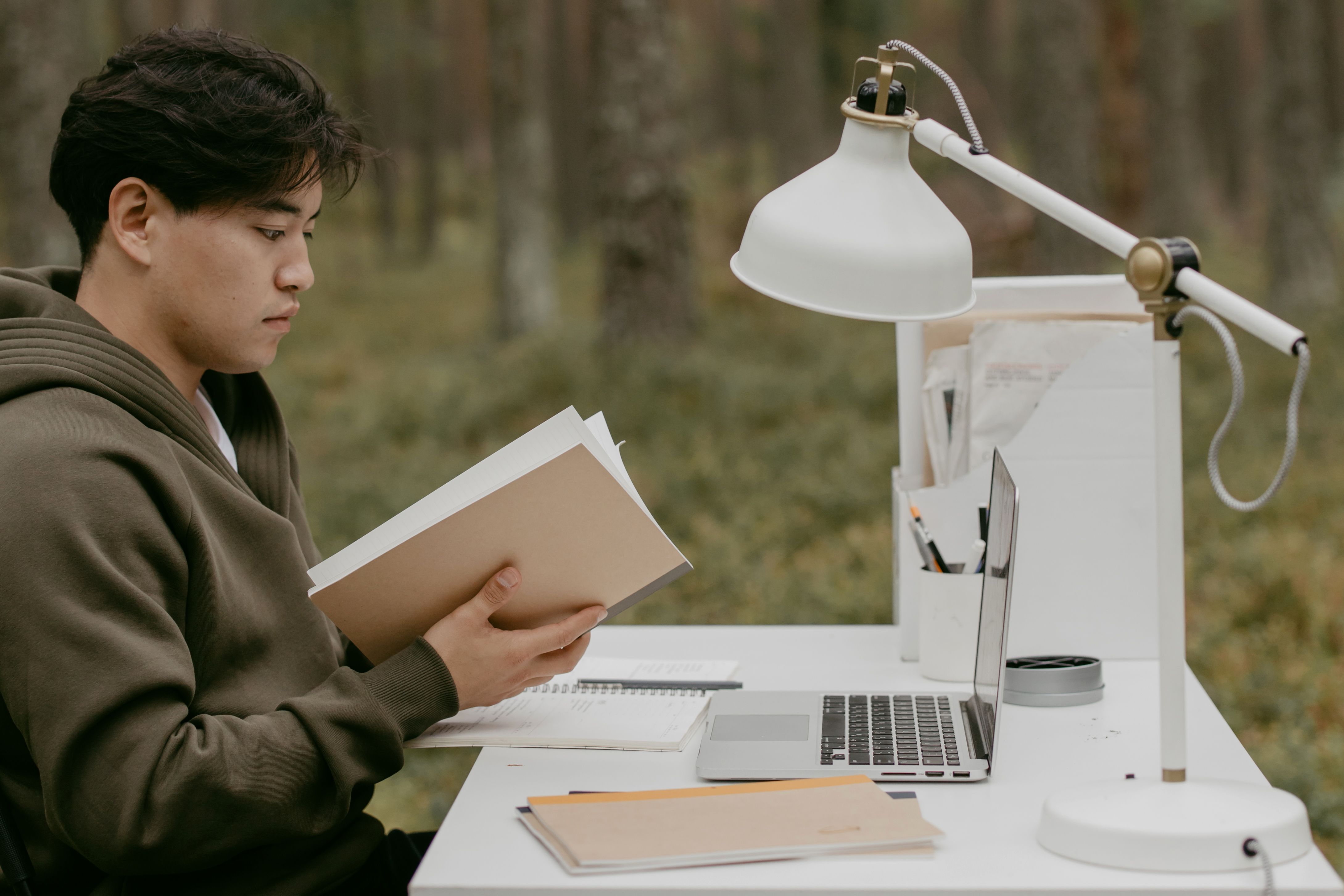 Asian man reading at an outdoor workspace with laptop, books, and lamp on desk.